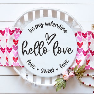 May include: White round wooden sign with the text "be my valentine hello love love sweet love" in black script. The sign is sitting on a white tray with a white background.