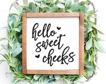 May include: A wooden sign with the text "hello sweet cheeks" in black script font. The sign is surrounded by green foliage and is sitting in a white wicker basket.