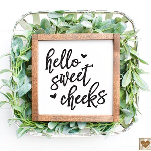 May include: A wooden sign with the text "hello sweet cheeks" in black script font. The sign is surrounded by green foliage and is sitting in a white wicker basket.