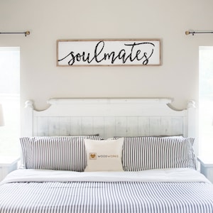 May include: A white bedroom with a white bed with a black and white striped comforter and pillows. There are two white nightstands with lamps on each side of the bed. The headboard has a sign above it that says "soulmates".