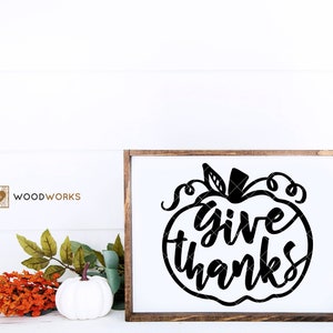 Puede incluir: Un letrero de madera con marco marrón y fondo blanco. El letrero presenta una silueta negra de una calabaza con el texto "I give thanks" en su interior.