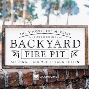 Puede incluir: Un letrero de madera blanco con texto negro que dice "The S'more, The Merrier. Let's Get Toasted. Backyard Fire Pit. Sit Long, Talk Much, Laugh Often." El letrero está enmarcado en madera oscura y está sentado en una pared de ladrillo.