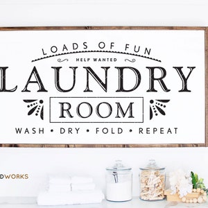 May include: A white wooden sign with black text that reads "Loads of Fun Help Wanted Laundry Room Wash Dry Fold Repeat". The sign is hanging on a white wall with a wooden frame.