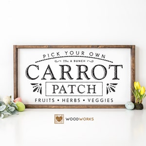 Op de afbeelding: Een houten bord met zwarte tekst die "Pick Your Own 25c a Bunch Carrot Patch Fruits Herbs Veggies Woodworks" zegt. Het bord is ingelijst in bruin hout en staat op een wit oppervlak met een groene bladertak en gele bloemen.