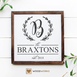 May include: A white wooden sign with a brown frame, featuring a black and white design. The sign reads "The Braxtons est. 2018" with a stylised letter "B" surrounded by a wreath.