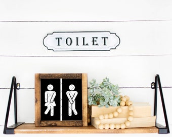 May include: A wooden sign with a black background and white stick figures depicting a woman and a man. The sign is on a shelf with a metal sign that reads "TOILET" above it.