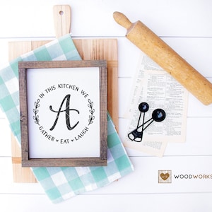 May include: A wooden sign with a brown frame and a white background. The sign says "In this kitchen we gather eat laugh" with a decorative wreath and the letter "A" in the center. The sign is sitting on a wooden cutting board with a green and white checkered cloth.