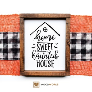 May include: A wooden sign with the text "Home Sweet Haunted House" in black script font. The sign is framed in a dark brown wood frame and has a white background. The sign is decorated with a small house shape and bats.