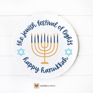 May include: A white round wooden sign with the text "The Jewish Festival of Lights Happy Hanukkah" in blue and gold lettering. A gold menorah with blue candles is in the center of the sign. Two blue Star of David symbols are on either side of the menorah.