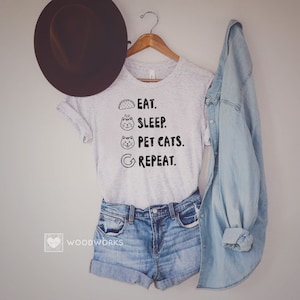 May include: A grey t-shirt with the text "Eat. Sleep. Pet Cats. Repeat." printed on it. The text is in black and includes a taco, a cat sleeping, a cat with its tongue out, and a circular arrow.
