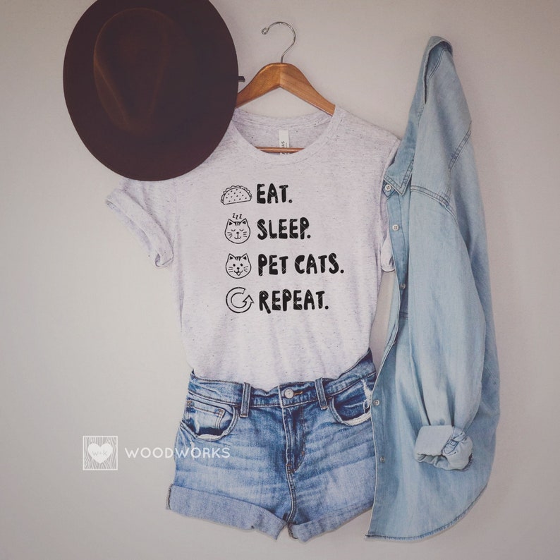 May include: A grey t-shirt with the text "Eat. Sleep. Pet Cats. Repeat." printed on it. The text is in black and includes a taco, a cat sleeping, a cat with its tongue out, and a circular arrow.