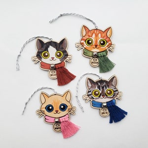 Może przedstawiać: Cztery drewniane ozdoby w kształcie kota z kolorowymi szalikami i frędzlami. Każdy kot ma unikalny wzór i plakietkę z imieniem: "Mojito", "Moscow", "Mimosa" i "Mai Tai". Są nawleczone na srebrno-biały sznurek, gotowe do zawieszenia.