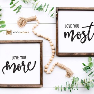 May include: Two wooden framed signs with white backgrounds and black calligraphy. The top sign reads "Love You Most" and the bottom sign reads "Love You More".