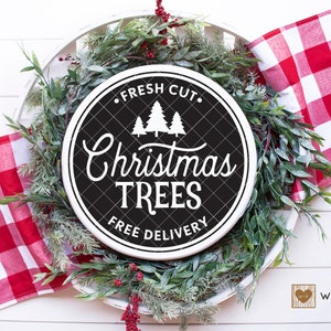 May include: A white round sign with black lettering that says "Fresh Cut Christmas Trees Free Delivery". The sign is surrounded by a green wreath with red berries.