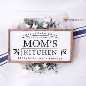 May include: A white wooden sign with a brown frame reads "Love Served Daily Open 24 Hours Mom's Kitchen Breakfast Lunch Dinner".