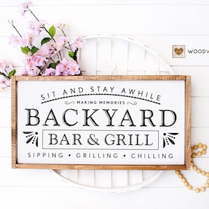 Może przedstawiać: Drewniany znak z napisem "Sit and Stay Awhile Making Memories Backyard Bar & Grill Sipping Grilling Chilling" czarnymi literami na białym tle. Znak ma brązową drewnianą ramę.