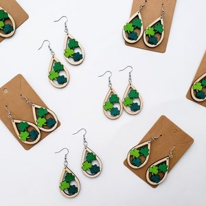 May include: Wooden teardrop earrings with a light wood frame and three green shamrocks inside. The shamrocks are in shades of green. The earrings hang from silver-colored hooks. Several pairs of earrings are displayed on brown cardboard earring cards.