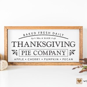 May include: A rustic wooden sign with the text "BAKED FRESH DAILY 50¢ A SLICE THANKSGIVING PIE COMPANY APPLE CHERRY PUMPKIN PECAN" in black lettering. The sign is on a white surface with two small pumpkins.