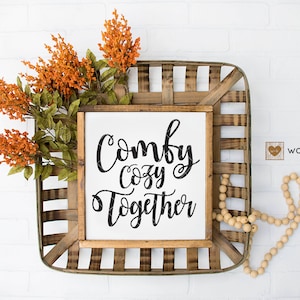 以下が含まれることがあります： 黒い筆記体で「Comfy Cozy Together」と書かれた木製の看板。看板は、木製のビーズの糸が置かれた籐のバスケットに入っています。