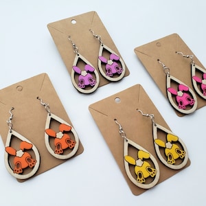 May include: Four pairs of wooden dangle earrings with a bunny rabbit silhouette. The earrings are in pink, orange, and yellow.