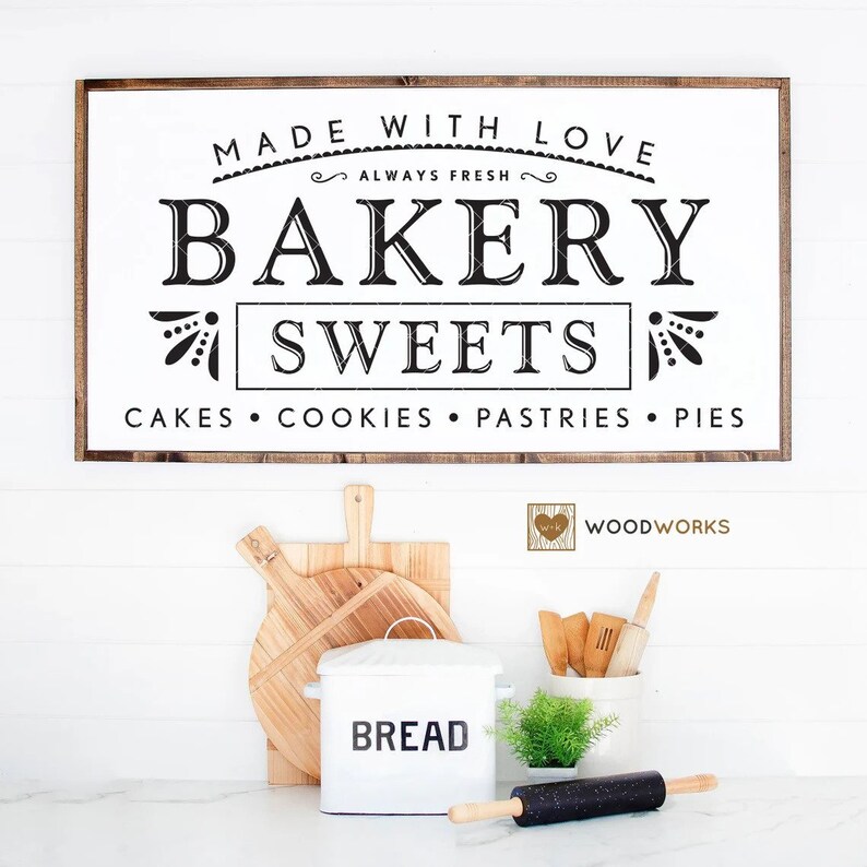 May include: A white wooden sign with brown trim reads "Made with Love Always Fresh Bakery Sweets Cakes Cookies Pastries Pies" in black text. The sign is hanging on a white wall above a kitchen counter with a white bread box, a wooden cutting board, and a rolling pin.