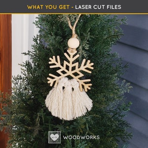 May include: A wooden snowflake ornament with a beige tassel hanging from a branch of a green artificial Christmas tree. The ornament is laser cut and has a wood grain pattern. The text "WOODWORKS" is visible in the bottom right corner of the image.