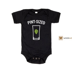 May include: Black baby onesie with white text that reads "PINT-SIZED" and a graphic of a pint glass with a green hop inside. The onesie has a white heart with the text "WOODWORKS" in brown.