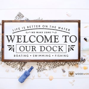 Puede incluir: Un letrero de madera con el texto "Life is better on the water, No Wake Zone, Welcome to Our Dock, Boating, Swimming, Fishing" en letras negras. El letrero tiene un marco de madera marrón y está rodeado de conchas y una red de pesca.