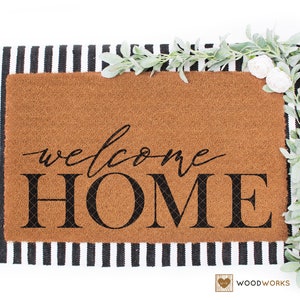 May include: A brown coir doormat with the text "welcome HOME" in black lettering. The doormat is on top of a black and white striped rug. There are white flowers and green leaves on the right side of the image.