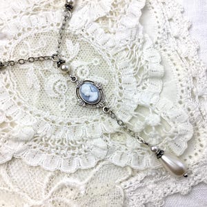 May include: A silver-toned necklace with a cameo pendant featuring a light blue profile. The necklace has a delicate chain design with pearl accents and a teardrop-shaped pearl drop. The necklace is displayed on a white lace doily.