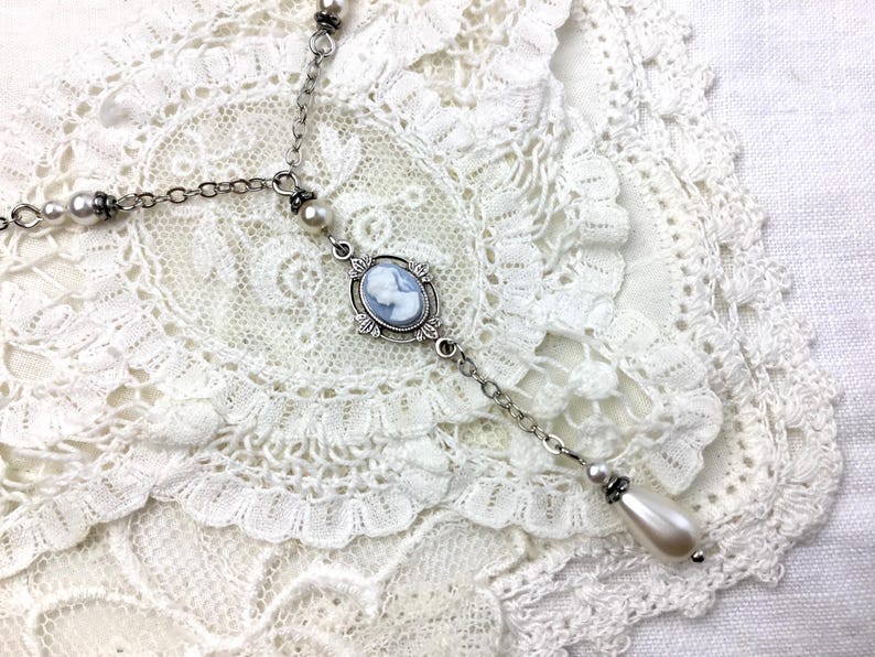 May include: A silver-toned necklace with a cameo pendant featuring a light blue profile. The necklace has a delicate chain design with pearl accents and a teardrop-shaped pearl drop. The necklace is displayed on a white lace doily.