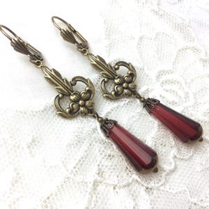 Op de afbeelding: Een paar oorhangers met een bronskleurig filigraanontwerp en een dieprode, traanvormige glazen kraal.