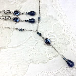 May include: A silver-toned jewelry set featuring a necklace and matching earrings. The necklace has a pendant with a dark blue oval stone and a teardrop-shaped dangle. The earrings also have dark blue oval stones and teardrop-shaped dangles. The jewelry is set on a white lace background.