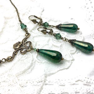 May include: An antique-style jewelry set featuring a necklace and matching earrings. The necklace has an ornate bronze-colored pendant with a floral design. The earrings and necklace feature faceted, teardrop-shaped green glass beads and smaller green crystal accents.