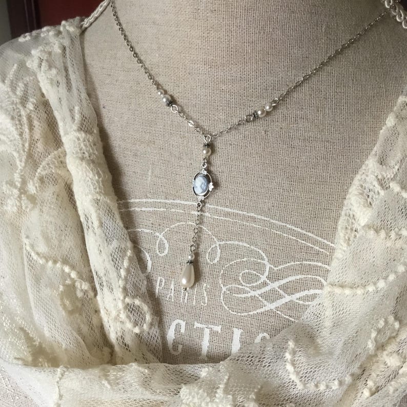 May include: A silver-toned necklace with a cameo pendant and a dangling pearl. The necklace features small pearls and a delicate chain. The cameo pendant has a blue and white design. The necklace is displayed on a mannequin.