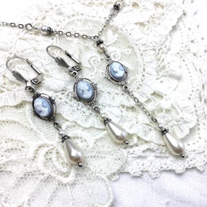 May include: A silver-toned jewelry set featuring a necklace and earrings. Each piece showcases a cameo pendant with a light blue profile, surrounded by an ornate frame. The earrings and necklace have dangling faux pearl accents, creating an elegant, vintage aesthetic. The set is displayed on a white lace surface.