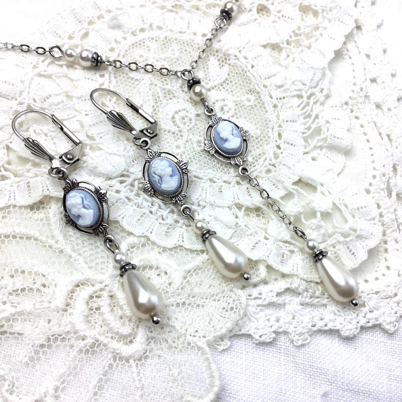 May include: A silver-toned jewelry set featuring a necklace and earrings. Each piece showcases a cameo pendant with a light blue profile, surrounded by an ornate frame. The earrings and necklace have dangling faux pearl accents, creating an elegant, vintage aesthetic. The set is displayed on a white lace surface.