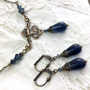 May include: A necklace and earring set featuring dark blue teardrop beads with ornate bronze-toned metal caps. The necklace includes a central decorative bronze filigree component and small faceted blue crystal accents along the chain.