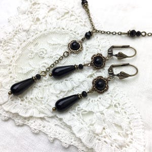 May include: A vintage jewelry set featuring a necklace and matching earrings. The set includes a delicate chain, black teardrop-shaped beads, and floral accents. The metal has a bronze finish, and the jewelry is displayed on a white lace surface.