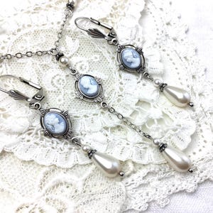 May include: Silver-toned jewelry set featuring cameo pendants and faux pearl accents. The set includes a necklace and two pairs of earrings. The earrings have a drop design with a pear-shaped pearl. The jewelry is displayed on a white lace surface.