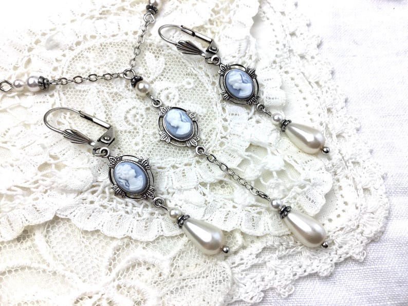 May include: Silver-toned jewelry set featuring cameo pendants and faux pearl accents. The set includes a necklace and two pairs of earrings. The earrings have a drop design with a pear-shaped pearl. The jewelry is displayed on a white lace surface.