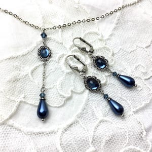 May include: A silver-toned jewelry set featuring a necklace and matching earrings. The necklace has a pendant with a dark blue oval stone and a dangling teardrop-shaped bead. The earrings have a similar design, with lever-back closures.