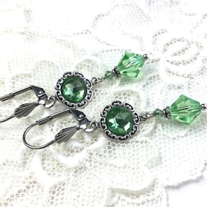 Op de afbeelding: Een paar oorhangers met lichtgroene facetkristallen en ronde groene stenen gezet in sierlijk zilverkleurig metaal. De oorbellen hebben oorhaaksluitingen en worden gepresenteerd op een wit kanten oppervlak.