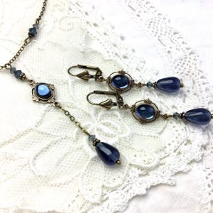 May include: A jewelry set consisting of a necklace and a pair of earrings crafted from antique bronze-toned metal. Each piece features oval blue cabochon stones set in ornate frames, accented with small blue crystal beads and teardrop-shaped dangles.
