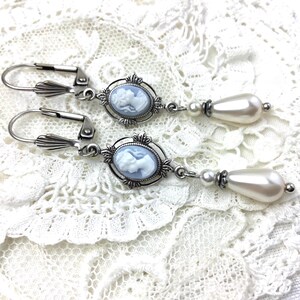 May include: A pair of silver-tone drop earrings. Each earring features a light blue cameo pendant with a white profile, surrounded by an ornate frame. Below the cameo hangs a large, pear-shaped faux pearl. The earrings rest on a white lace surface.