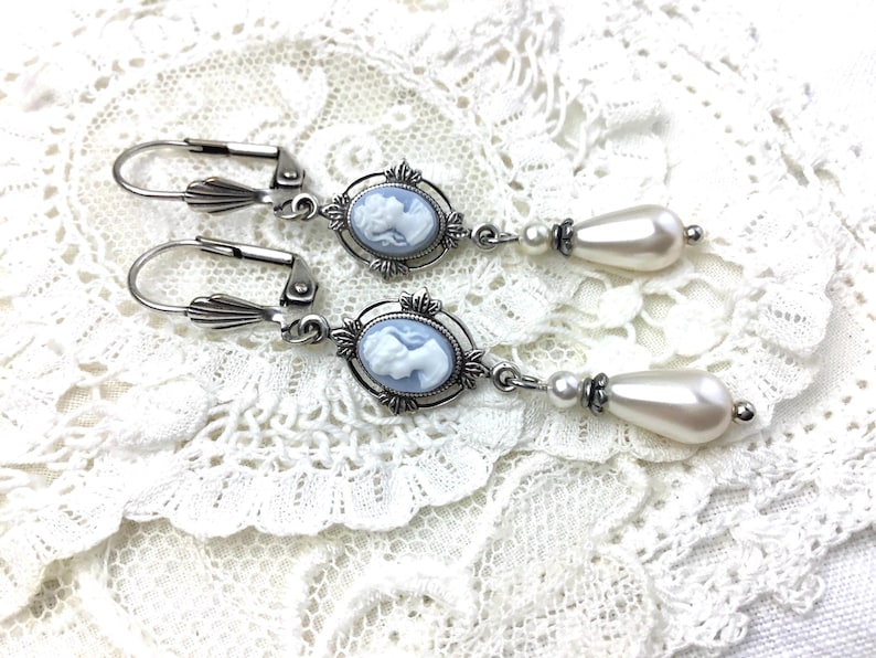 May include: A pair of silver-tone drop earrings. Each earring features a light blue cameo pendant with a white profile, surrounded by an ornate frame. Below the cameo hangs a large, pear-shaped faux pearl. The earrings rest on a white lace surface.