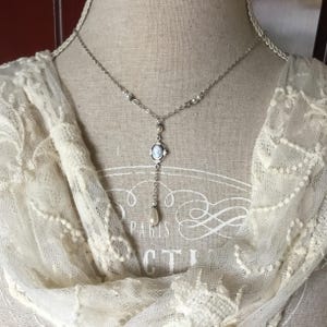 May include: Silver-toned pendant necklace with a cameo pendant and a dangling pearl. The necklace is displayed on a mannequin with a cream-colored lace scarf. The cameo features a profile image.