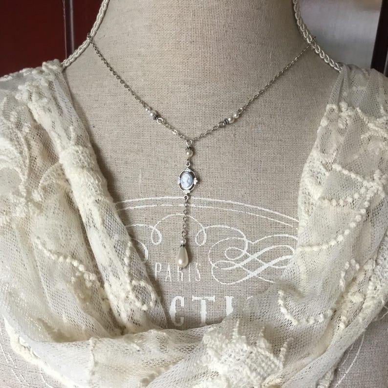 May include: Silver-toned pendant necklace with a cameo pendant and a dangling pearl. The necklace is displayed on a mannequin with a cream-colored lace scarf. The cameo features a profile image.