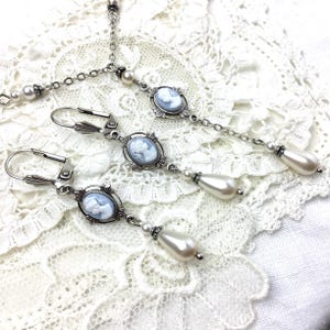May include: A vintage-style jewelry set featuring a necklace and earrings. The set includes a silver-toned chain necklace with a cameo pendant and pearl accents, and matching drop earrings with cameo pendants and teardrop-shaped faux pearls.