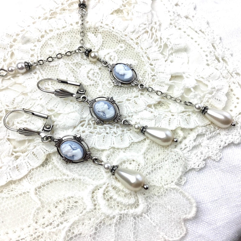 May include: A vintage-style jewelry set featuring a necklace and earrings. The set includes a silver-toned chain necklace with a cameo pendant and pearl accents, and matching drop earrings with cameo pendants and teardrop-shaped faux pearls.